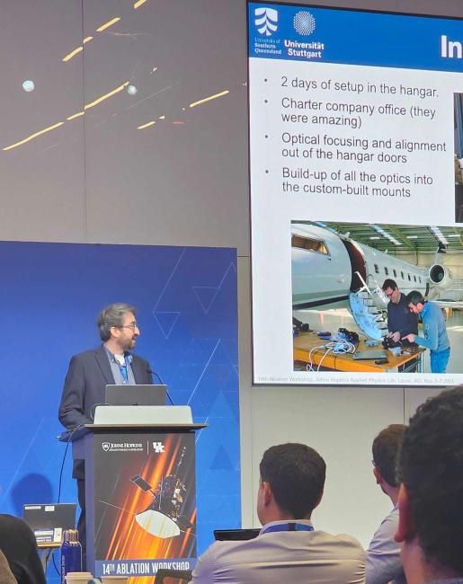 Speaker presenting at conference with projection screen showing research details